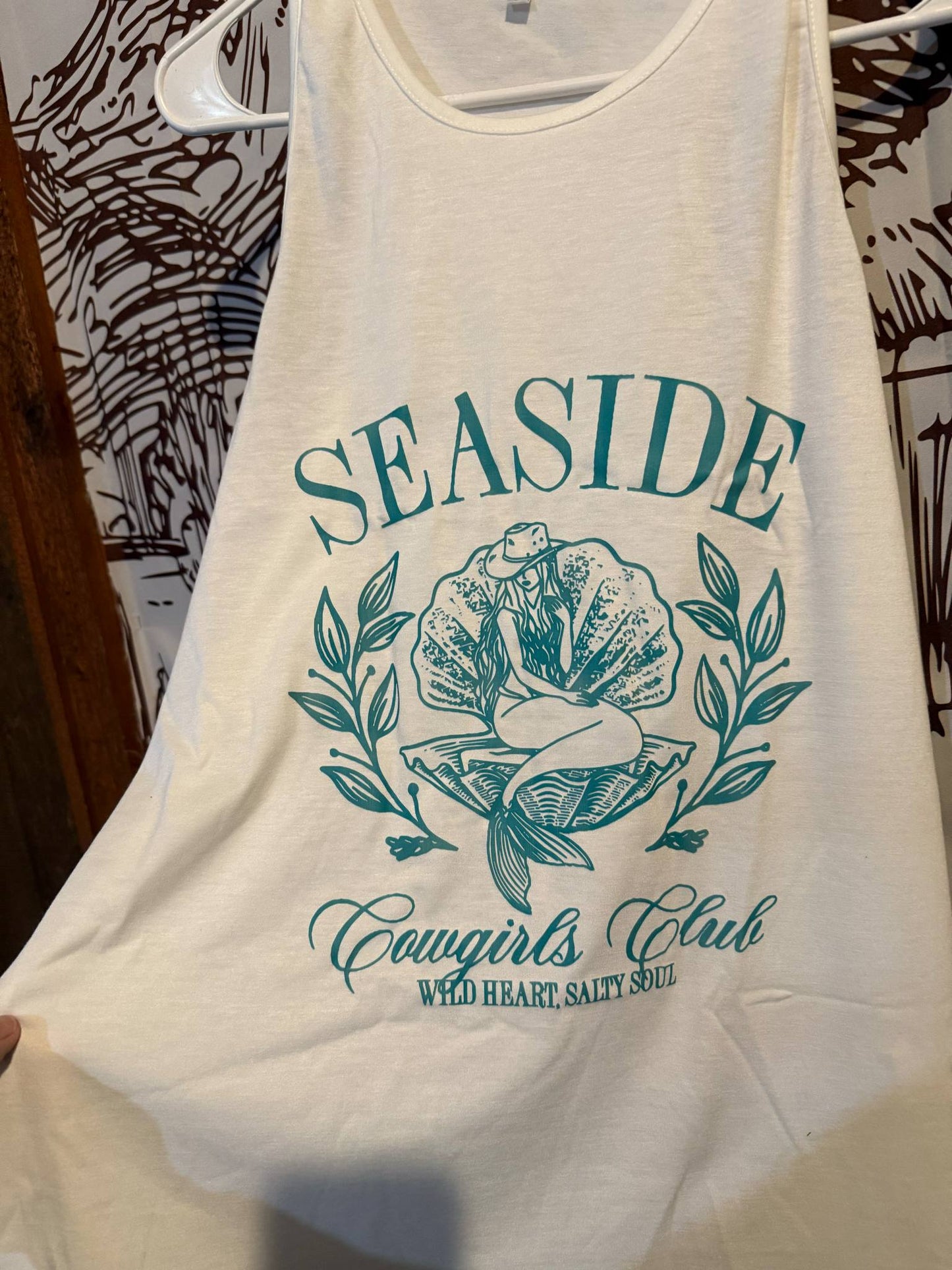 White tank top with teal design reading “Seaside Cowgirls Club – Wild Heart, Salty Soul” featuring a cowgirl-mermaid graphic surrounded by floral accents, displayed on a hanger.