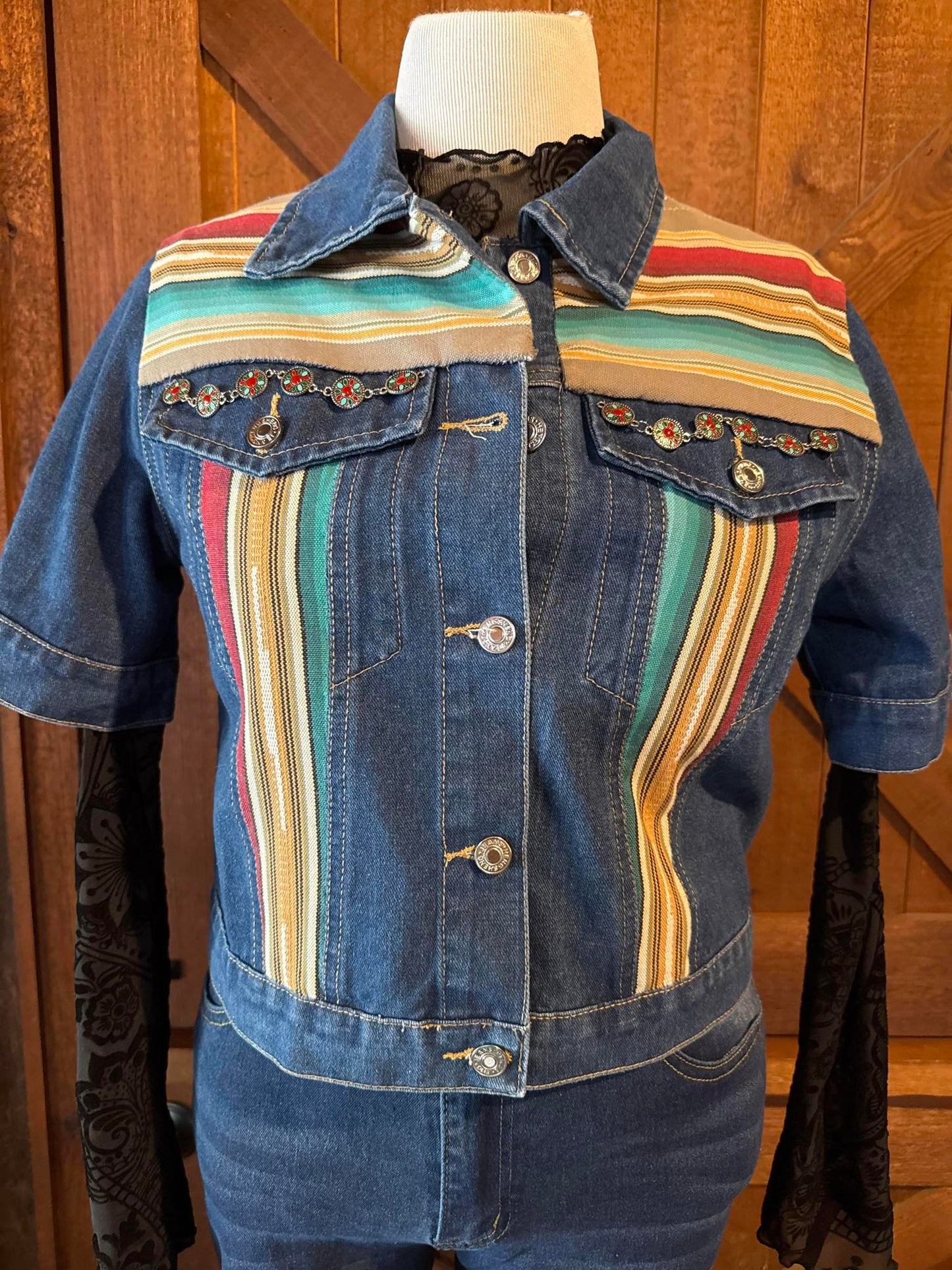 Short sleeve denim jacket with soft southwestern serape stripes in teal, tan, red, and gold, featuring decorative concho chain detail on chest pockets. Size Large and one of a kind.