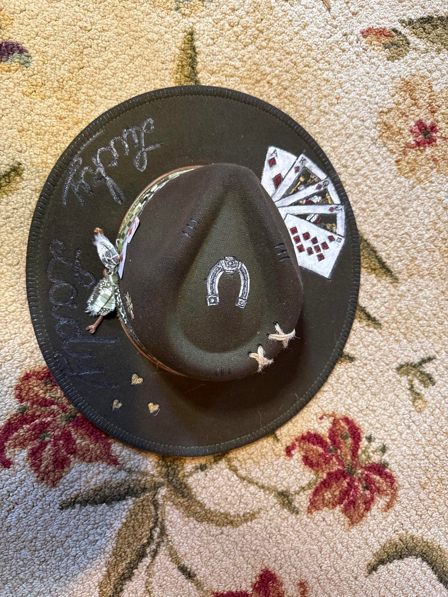 Black medium-brim fedora featuring embroidered playing cards, horseshoe, stitched stars, and script words like "lucky" on the brim, photographed on a floral rug.