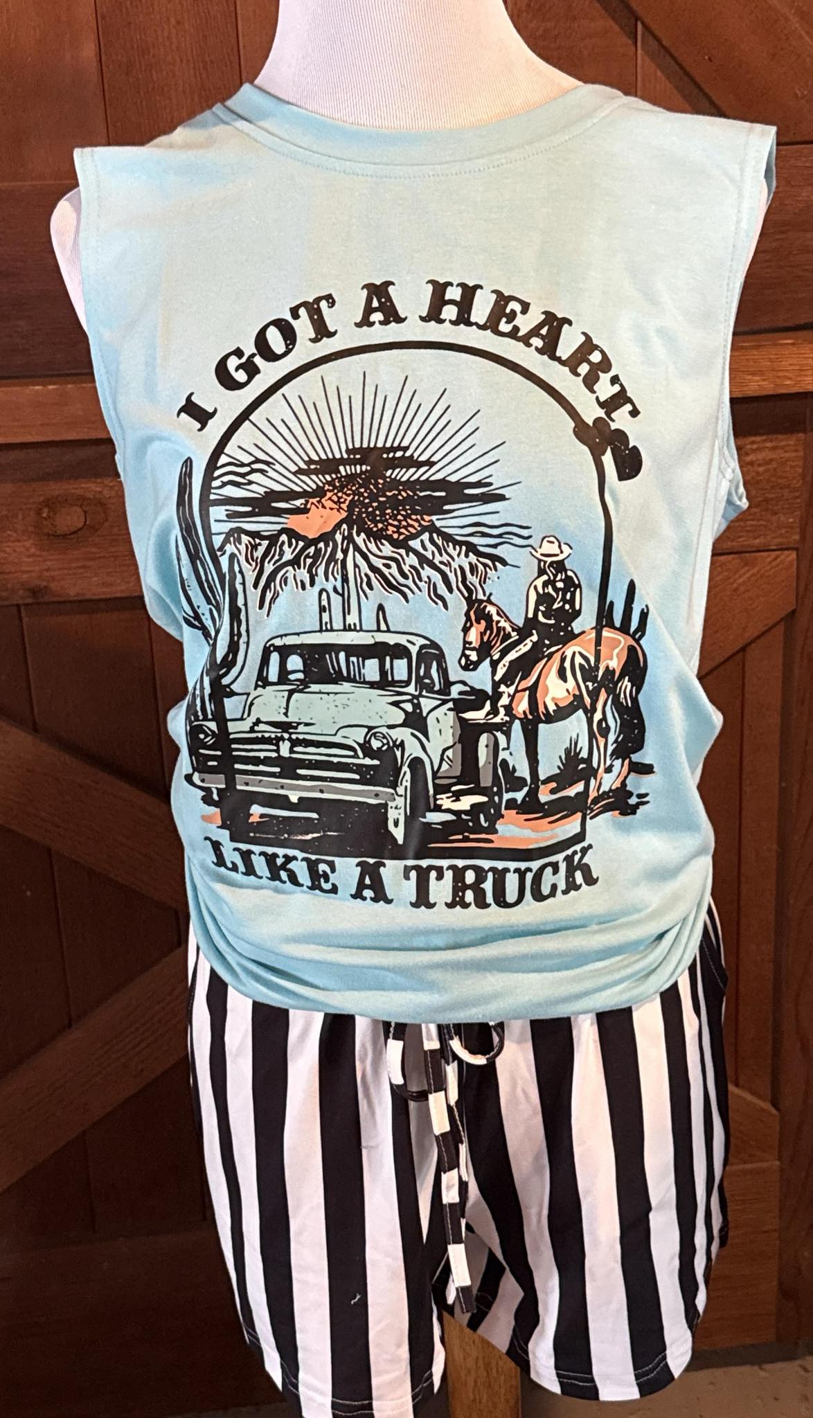 Light blue sleeveless tank top with a western graphic featuring a vintage pickup truck, a cowboy with a horse, a desert scene, and the phrase βI Got a Heart Like a Truck,β displayed on a mannequin with black and white striped shorts.