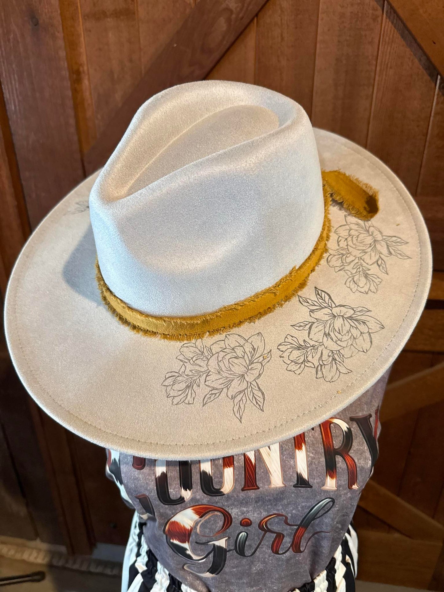 Soft cream-colored fedora hat with mustard yellow frayed band and delicate floral and hummingbird line art on the brim, displayed atop a western outfit.