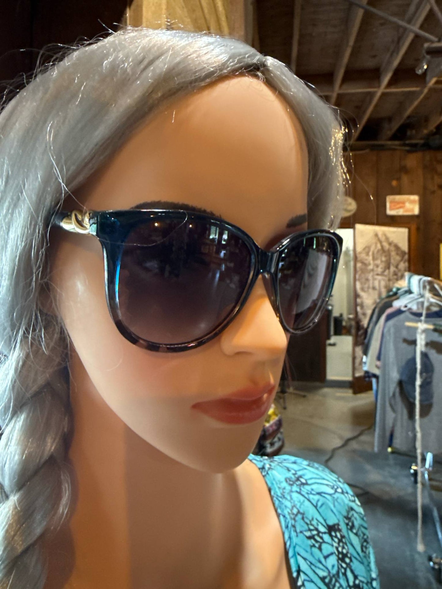 Mannequin wearing oversized black sunglasses with dark lenses and gold accents on the arms, styled with a silver braid and turquoise-patterned top inside a boutique.