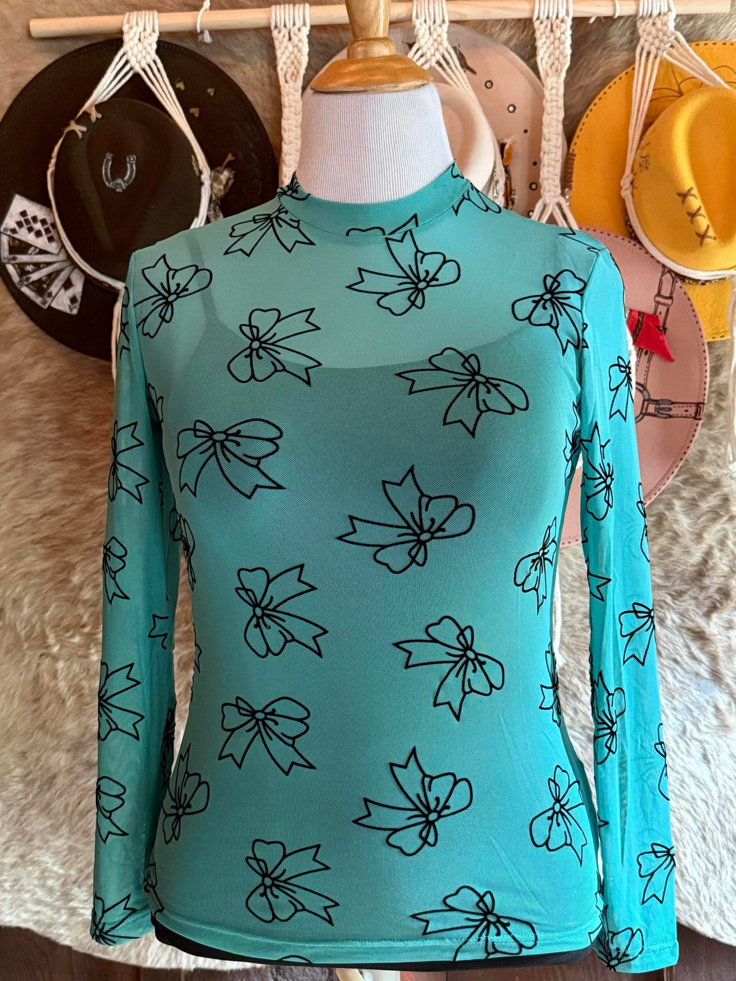Teal mesh long-sleeve top featuring black velvet bow designs, displayed on a mannequin with western hats in the background.