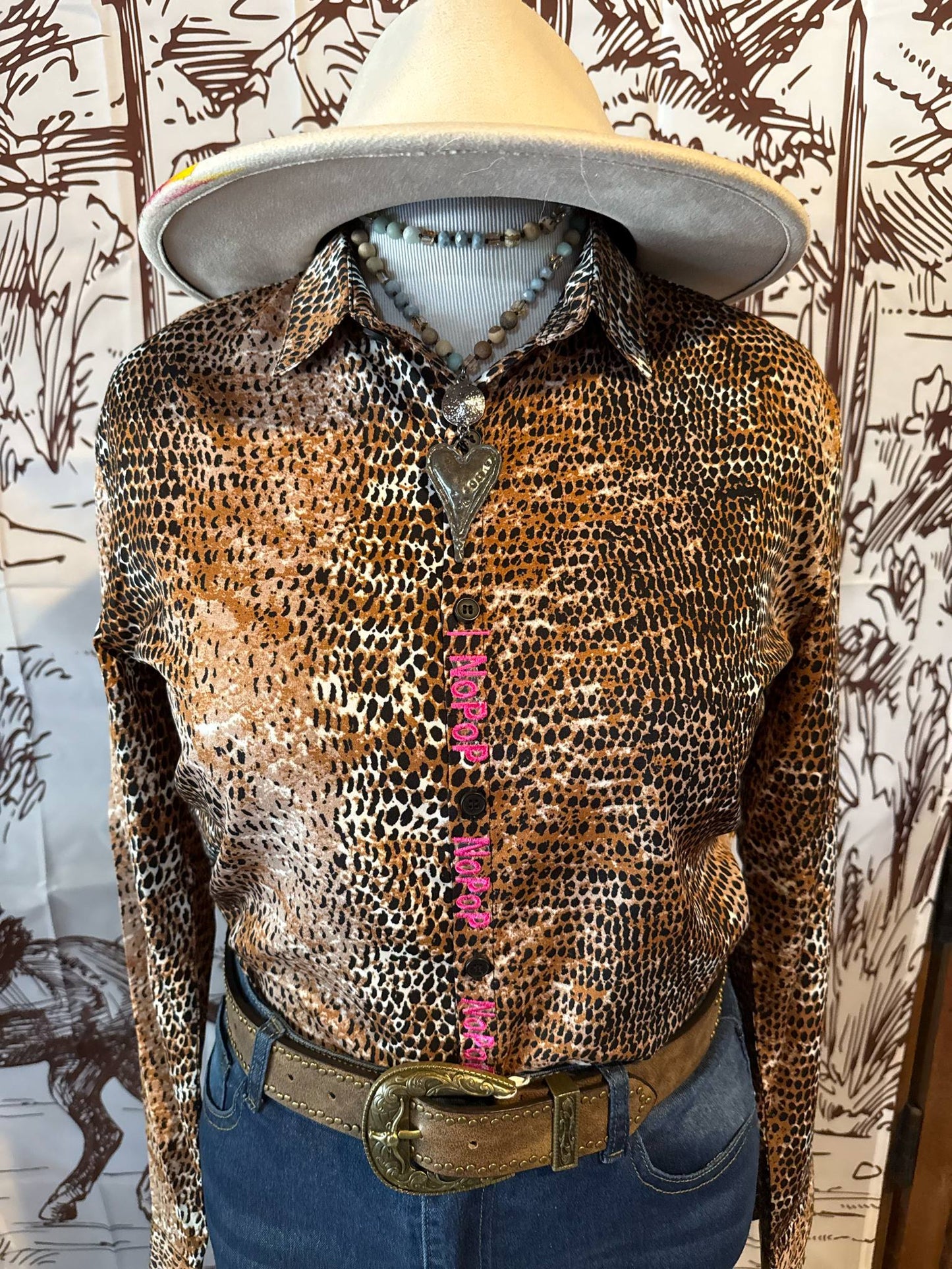 Western-style leopard print NoPop shirt with hot pink embroidered closure, shown on a mannequin styled with a beige cowboy hat, layered necklaces, and a tooled leather belt with jeans.