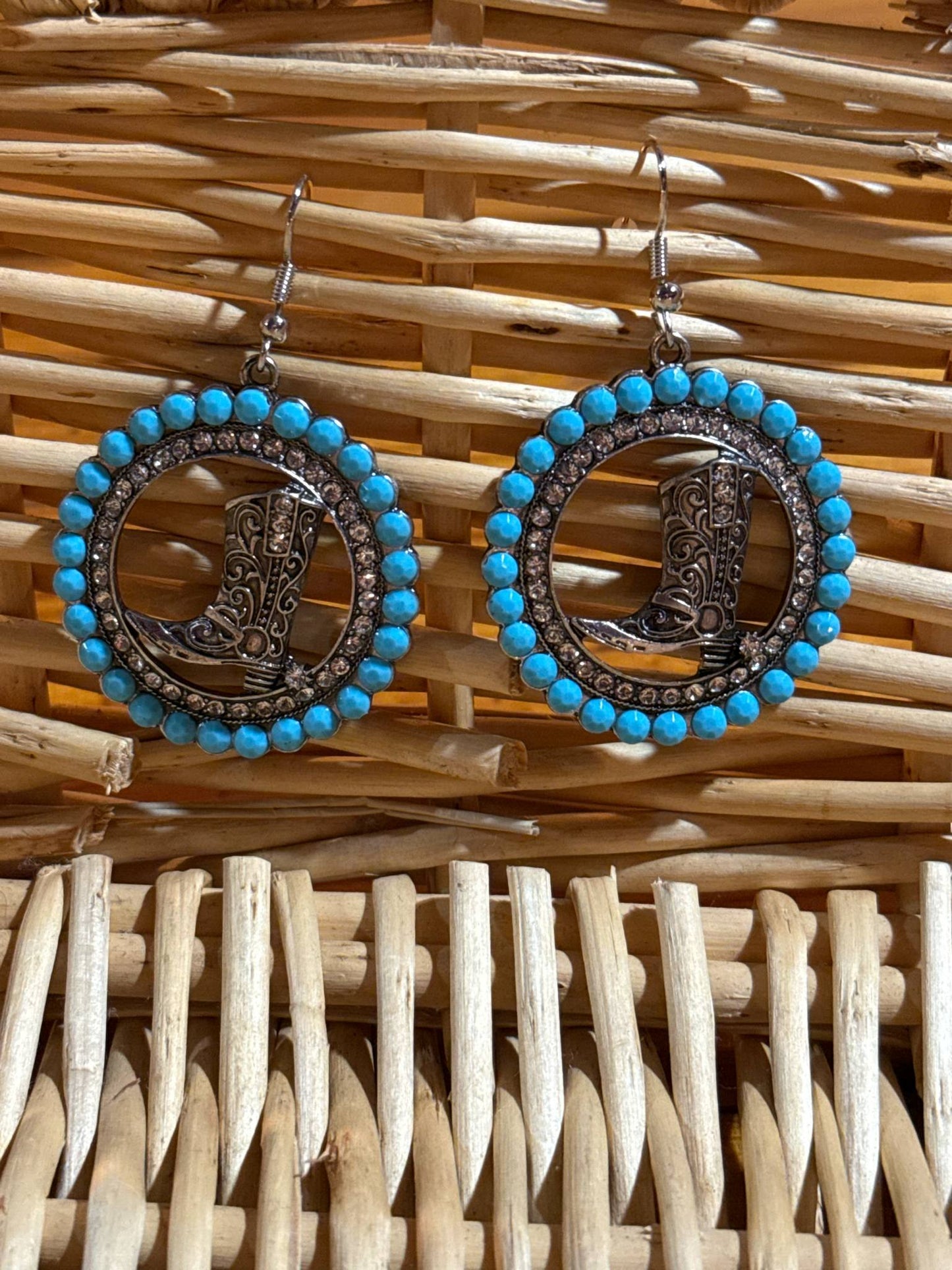 Pair of round silver dangle earrings with detailed cowboy boot cutouts in the center, surrounded by sparkling rhinestones and turquoise beading, displayed on a woven wicker background.
