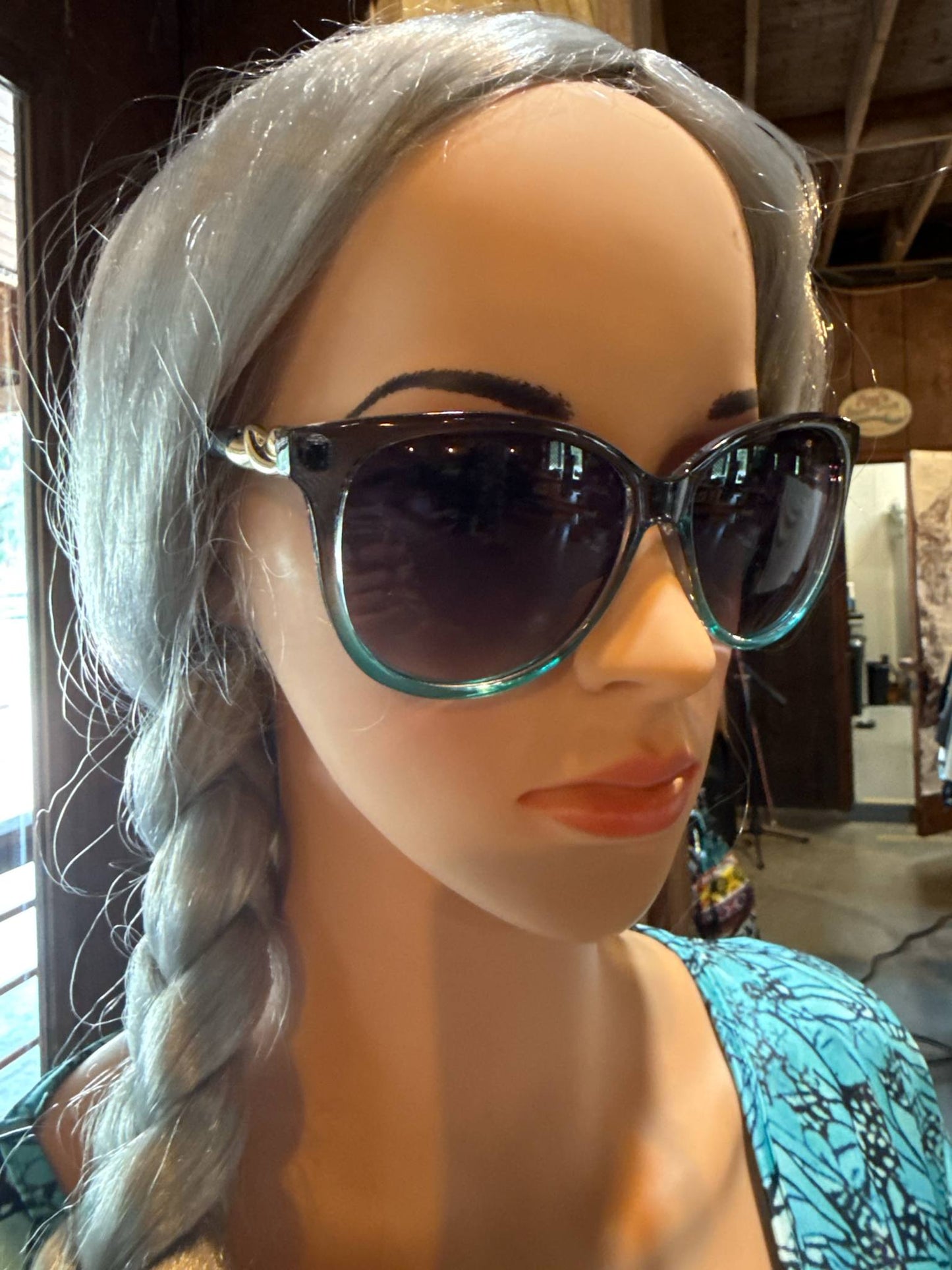 Mannequin wearing oversized sunglasses with a subtle teal blue frame and gold accents, styled with a side braid and turquoise-patterned top inside a boutique.