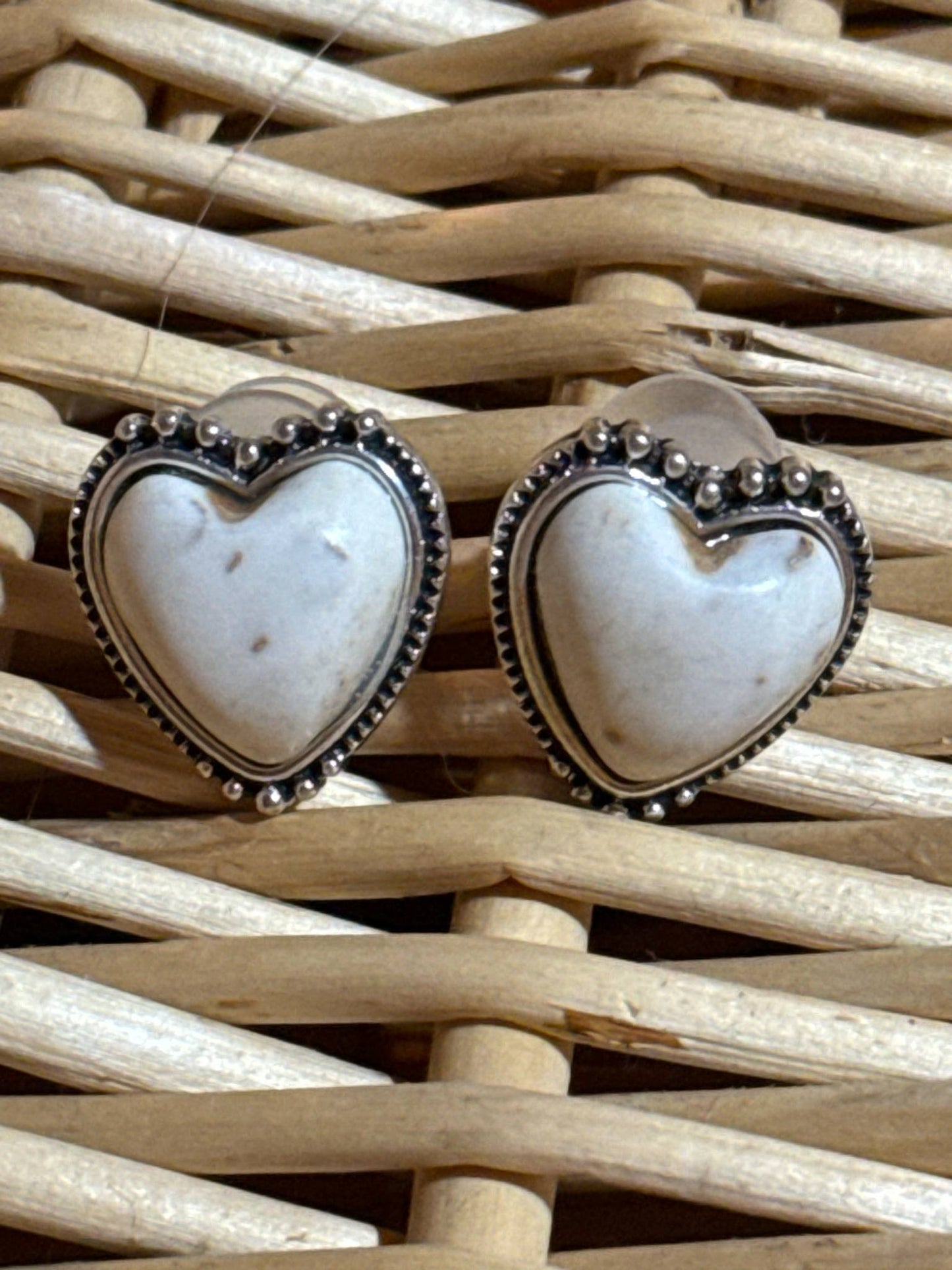 Pair of heart-shaped stud earrings with a white marbled stone center and silver beaded trim, displayed on a woven wicker background.