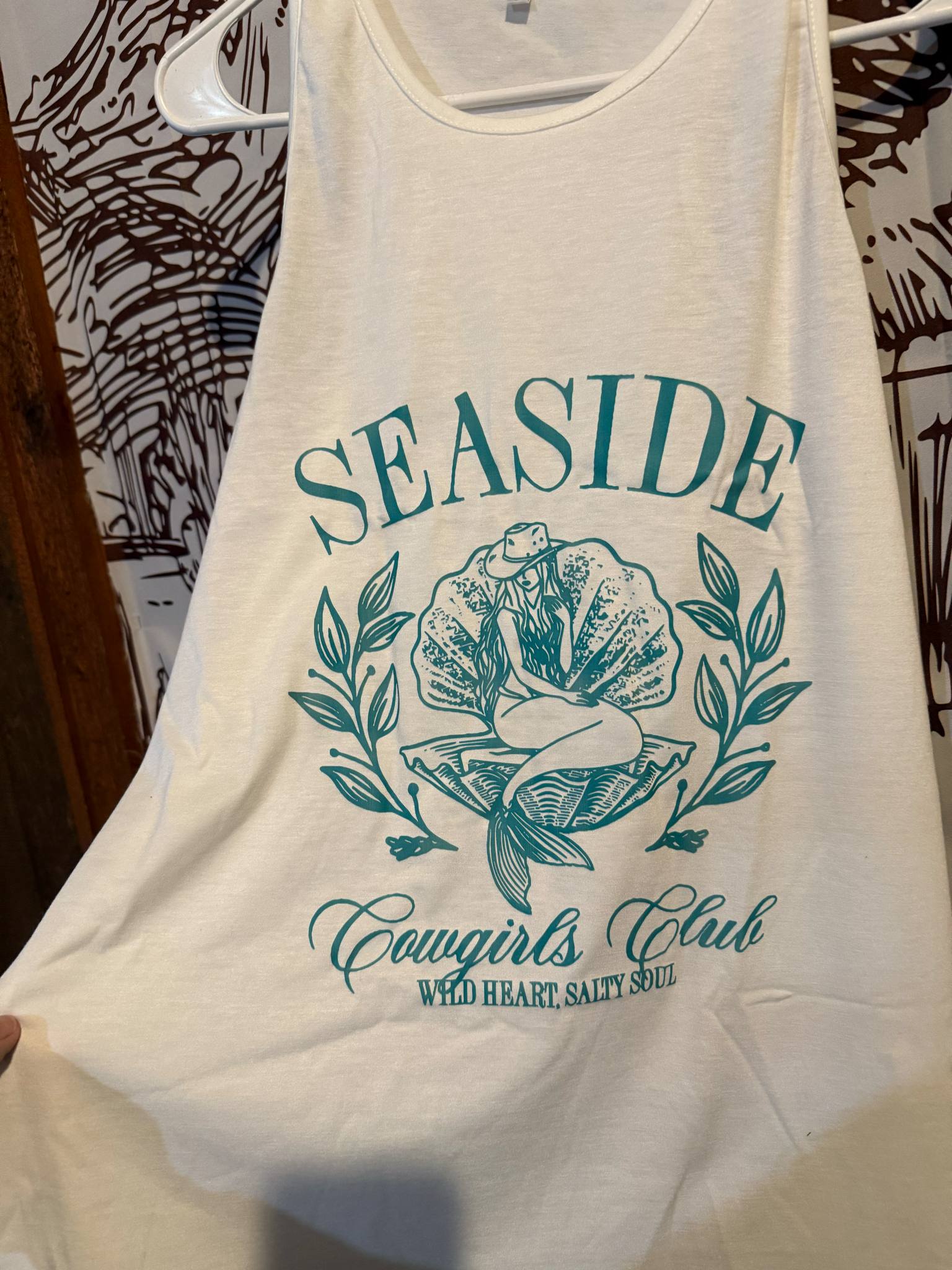 White tank top with teal design reading “Seaside Cowgirls Club – Wild Heart, Salty Soul” featuring a cowgirl-mermaid graphic surrounded by floral accents, displayed on a hanger.