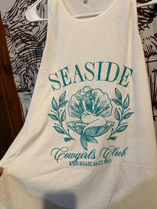 White tank top with teal design reading “Seaside Cowgirls Club – Wild Heart, Salty Soul” featuring a cowgirl-mermaid graphic surrounded by floral accents, displayed on a hanger.