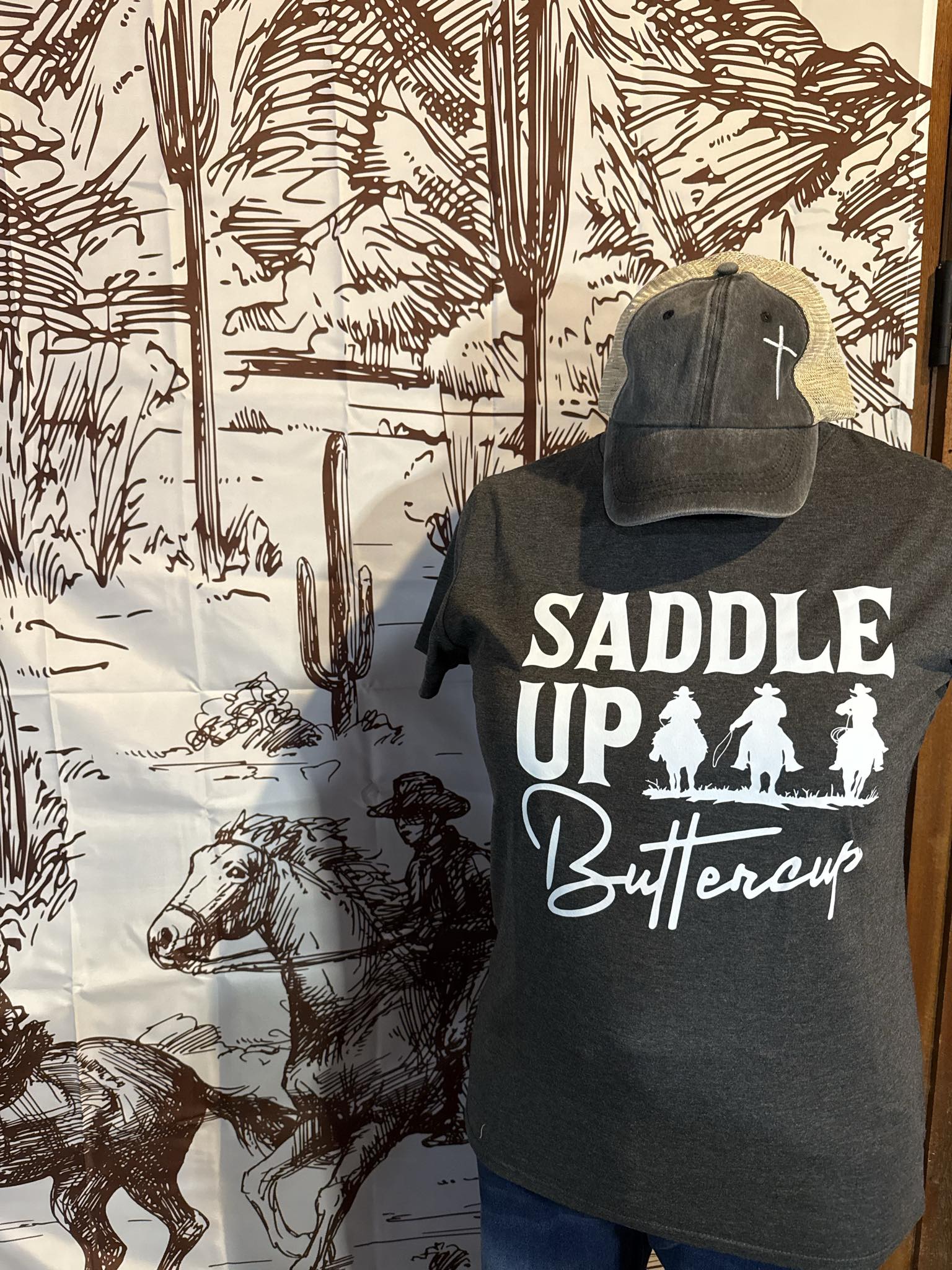Charcoal graphic tee with "Saddle Up Buttercup" text and cowboy silhouettes, paired with a distressed tan and charcoal mesh-back trucker hat featuring a small embroidered cross.