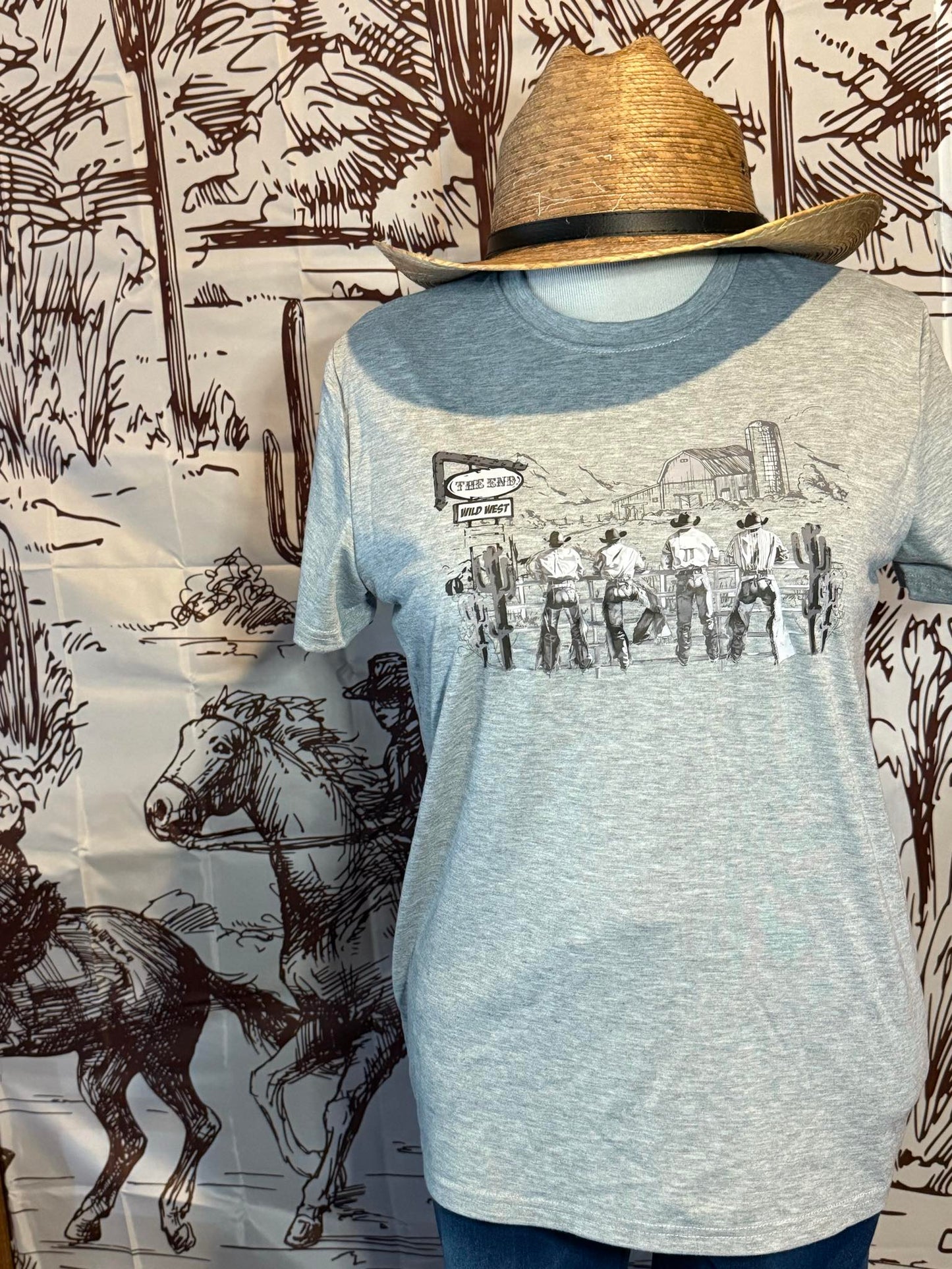 Light gray graphic tee featuring a Wild West cowboy scene paired with a palm leaf adjustable cowboy hat, styled on a mannequin with a Western-themed backdrop.