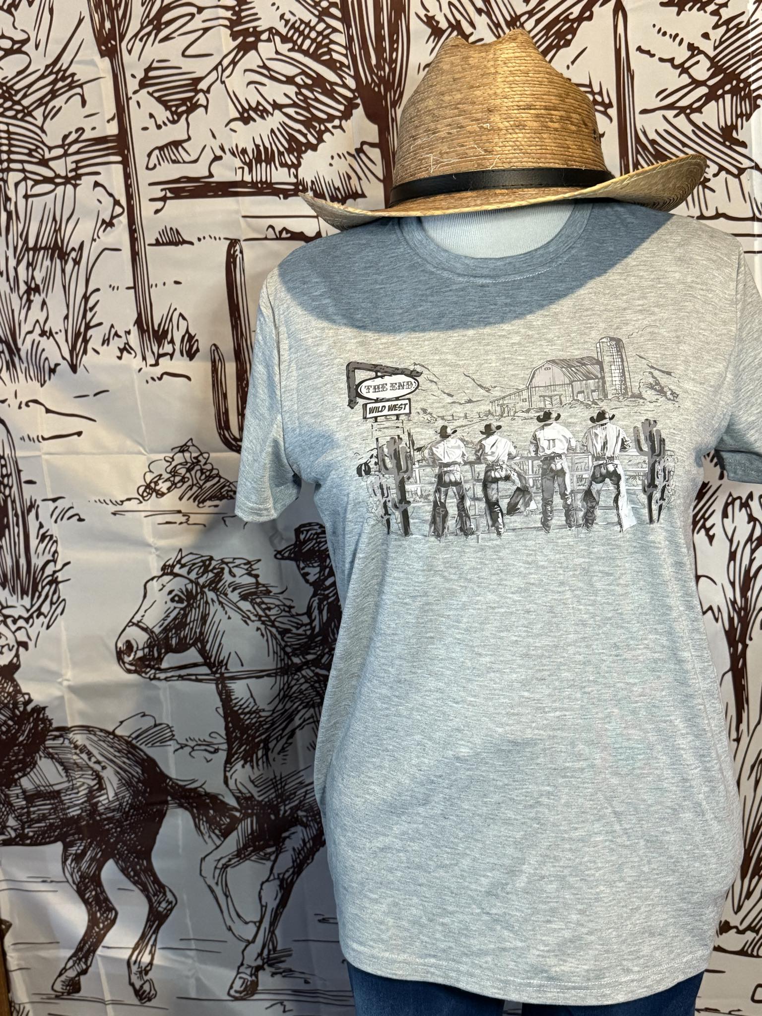Light gray graphic tee featuring a Wild West cowboy scene paired with a palm leaf adjustable cowboy hat, styled on a mannequin with a Western-themed backdrop.