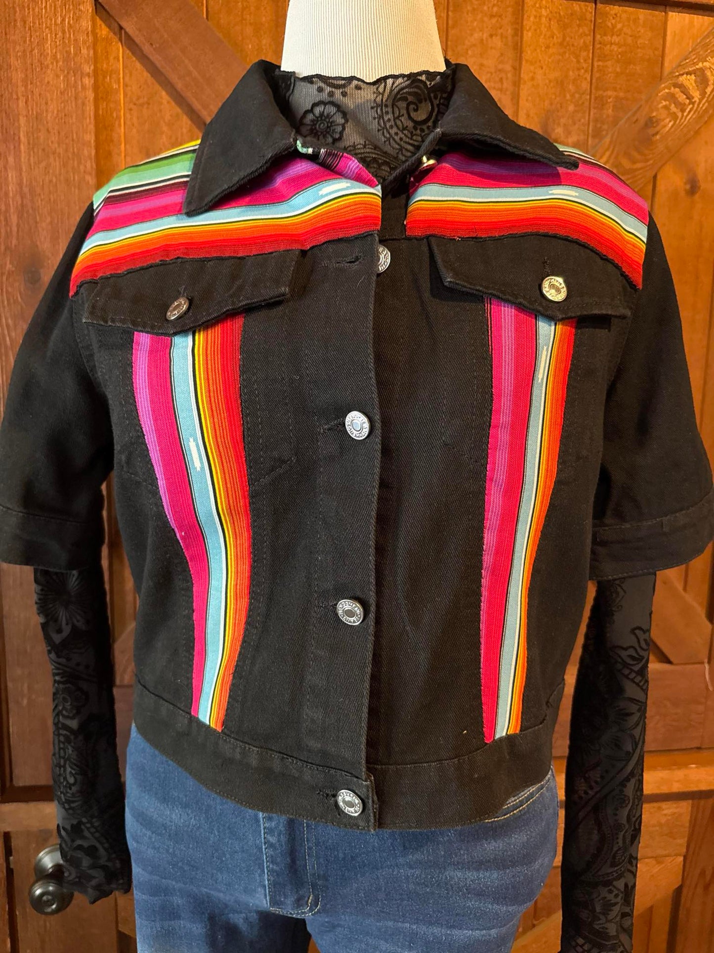 Hand-embellished black denim jacket featuring bright serape fabric in rainbow stripes on front and back, with short sleeves and silver snap buttons. Size Large.