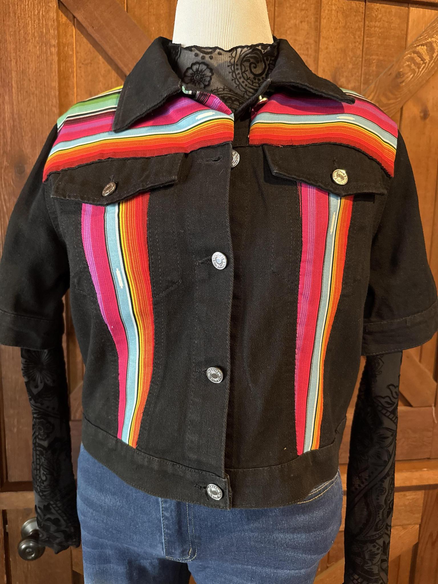 Hand-embellished black denim jacket featuring bright serape fabric in rainbow stripes on front and back, with short sleeves and silver snap buttons. Size Large.