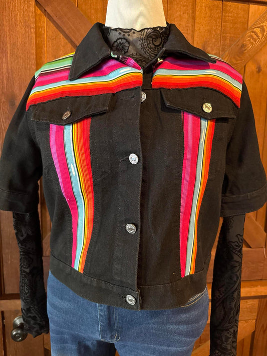 Hand-embellished black denim jacket featuring bright serape fabric in rainbow stripes on front and back, with short sleeves and silver snap buttons. Size Large.