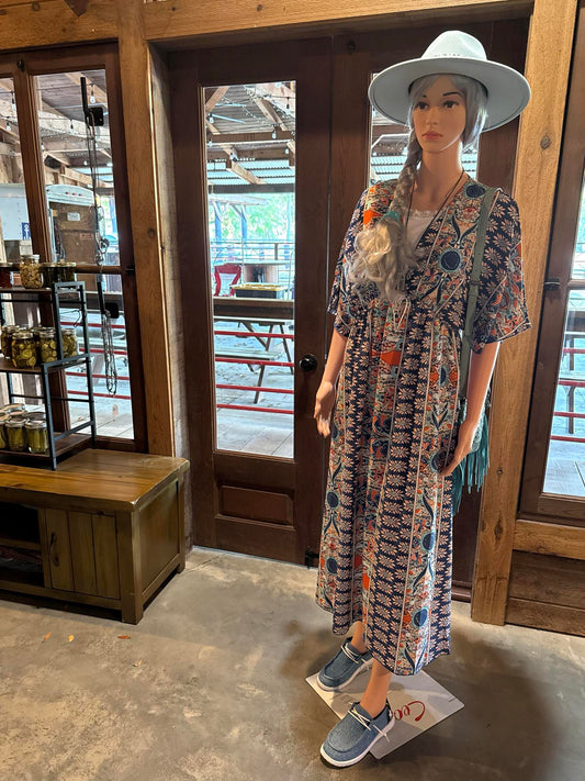 A flowy, boho-style summer dress with floral and paisley prints in blue, orange, and cream, displayed on a mannequin with a white hat and blue slip-on shoes.