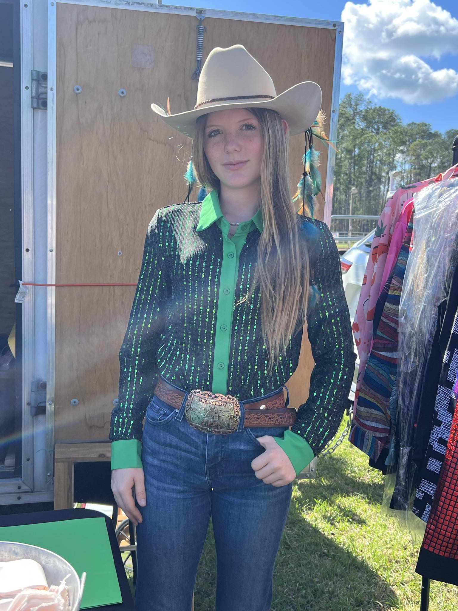 Cowgirl wearing a sheer, stretchy, lightweight green shimmer show shirt with jeans, a belt buckle, and a cowboy hat at an outdoor event.