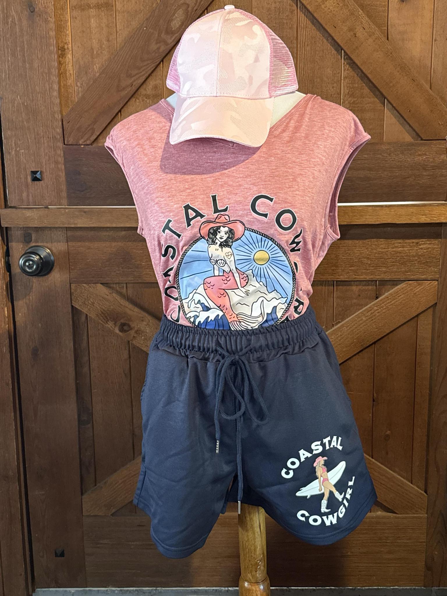 Navy blue drawstring shorts featuring a graphic of a cowgirl holding a surfboard with the text "Coastal Cowgirl" in white, styled on a mannequin with a matching tank top.