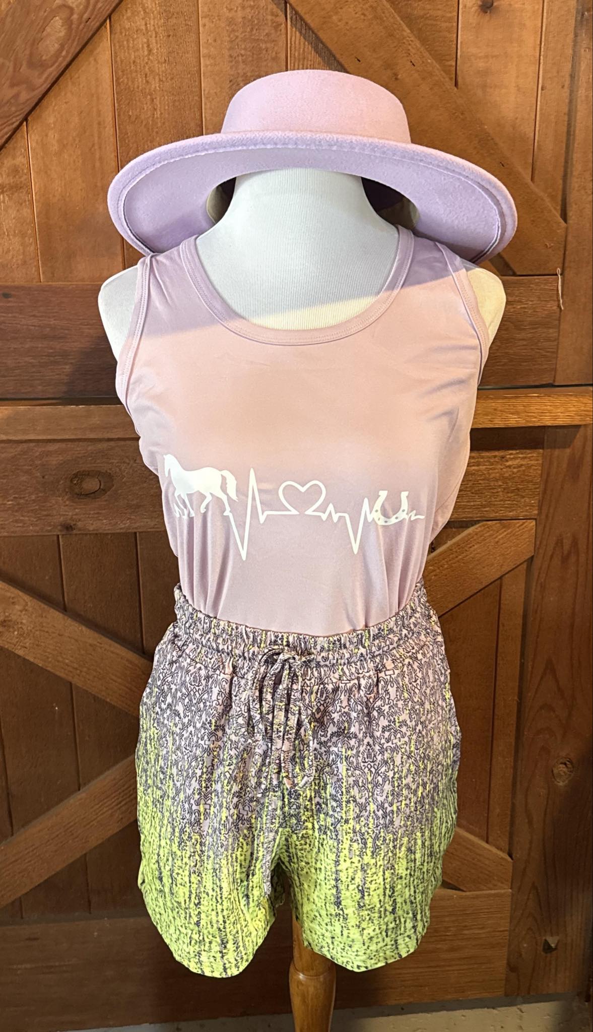 Lavender racerback tank top featuring a white graphic of a heartbeat line transforming into a horse, heart, and horseshoe, styled on a mannequin with lime ombré shorts and a wide-brim lavender hat.