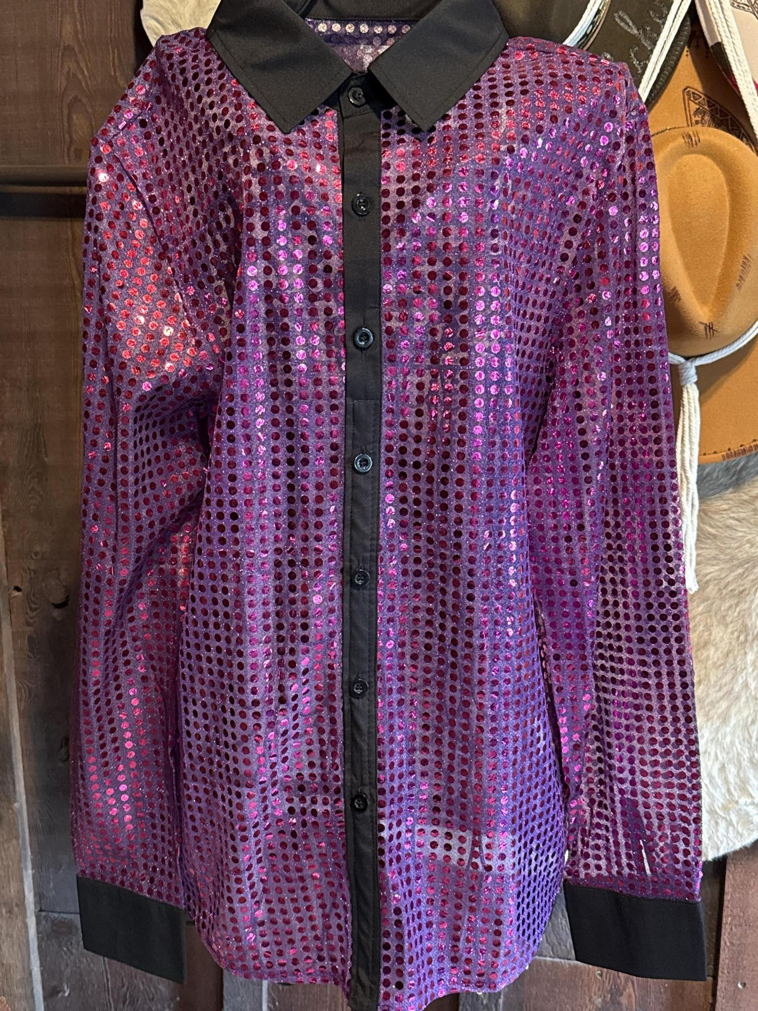 Sheer and shimmering long sleeve sequin button-up shirt in electric pink and purple hues with black cuffs and collar, hanging against a rustic wooden background.