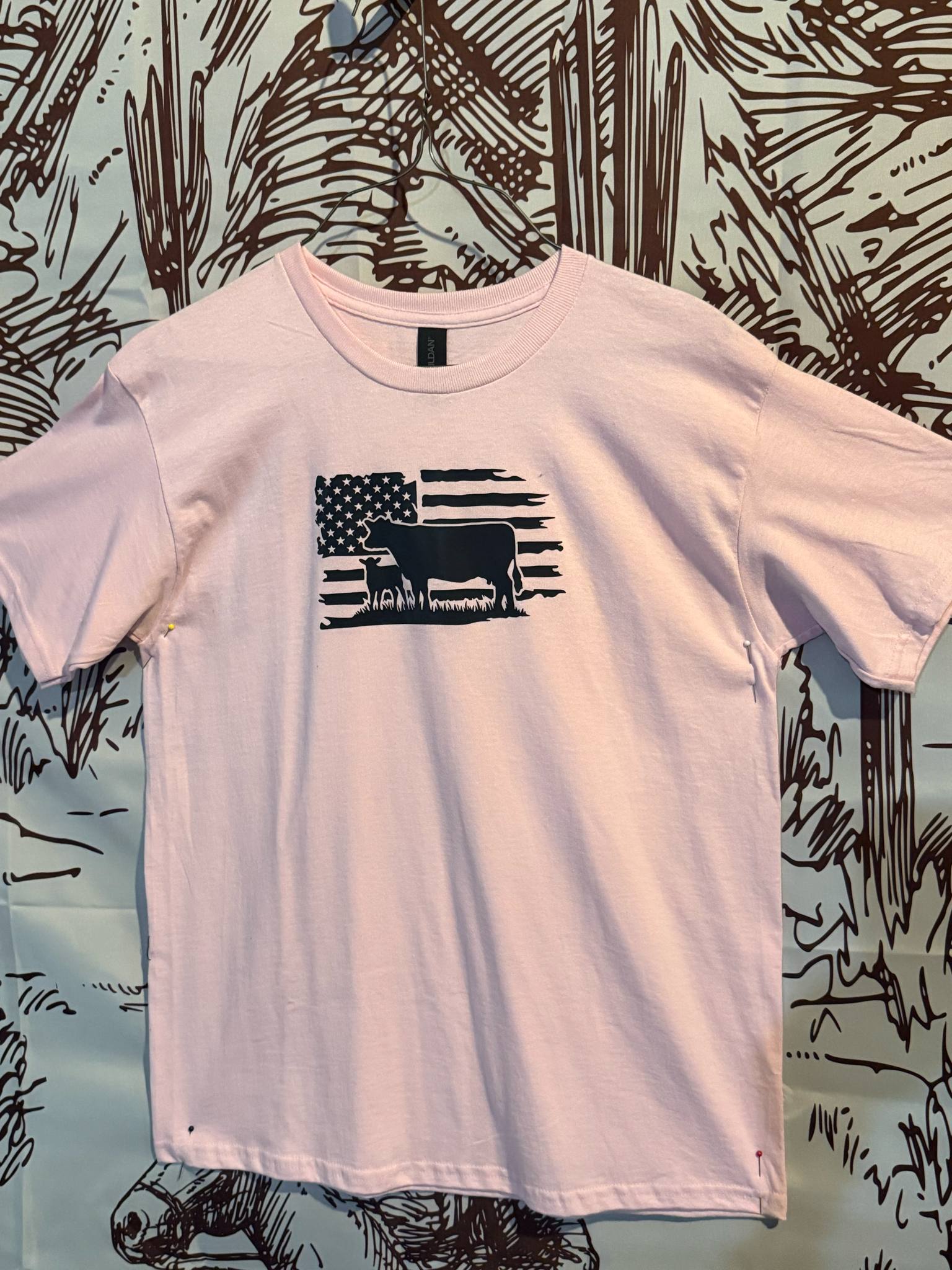 Light pink youth t-shirt featuring a black silhouette of a cow and calf in front of a distressed American flag graphic.