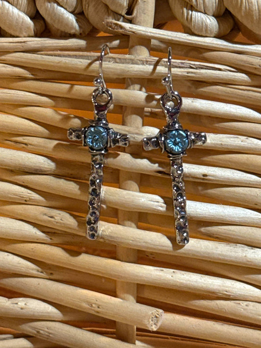A pair of silver-toned dangle earrings shaped like crosses, each with a turquoise floral center and small dark rhinestones lining the vertical bar, displayed against a woven wicker background.