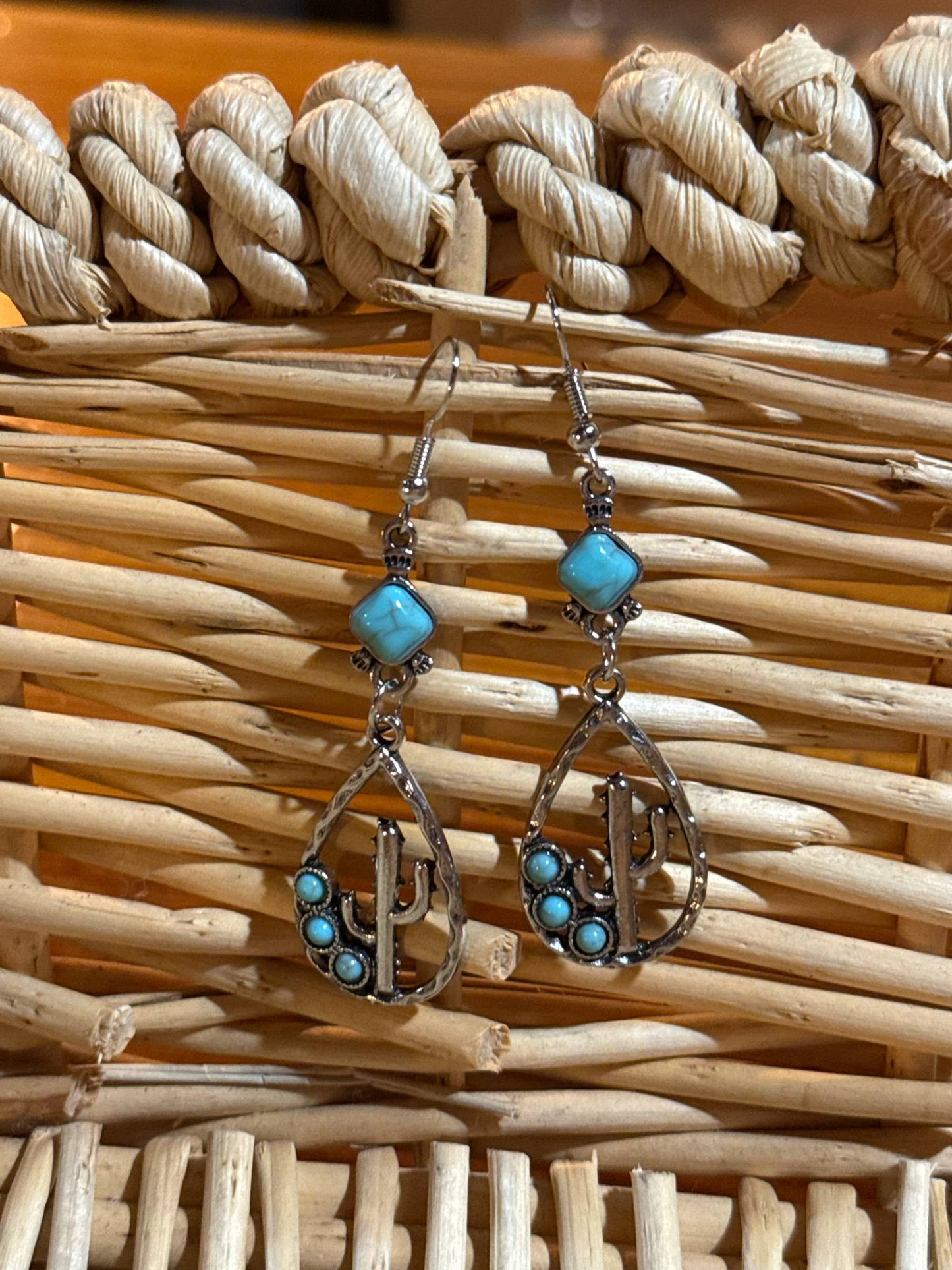 Pair of silver teardrop-shaped dangle earrings featuring a cactus design with small turquoise beads and a square turquoise stone at the top, displayed against a woven wicker background.