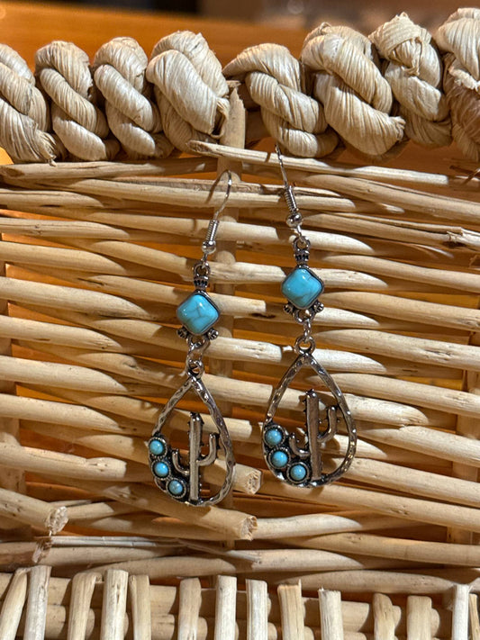 Pair of silver teardrop-shaped dangle earrings featuring a cactus design with small turquoise beads and a square turquoise stone at the top, displayed against a woven wicker background.
