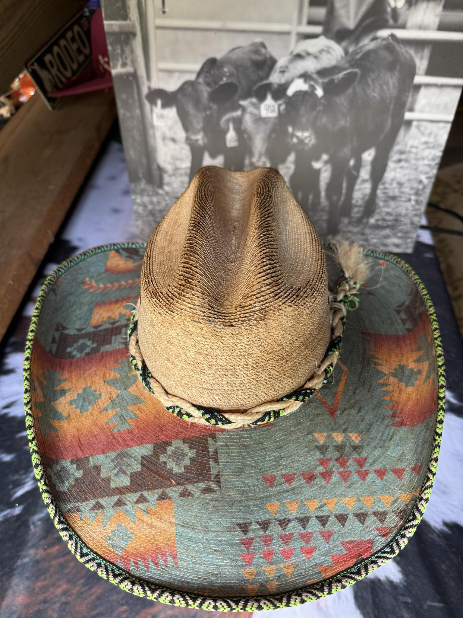 Natural straw cowboy hat with colorful Aztec-printed brim and braided hatband, shown from front and top view on a display stand.