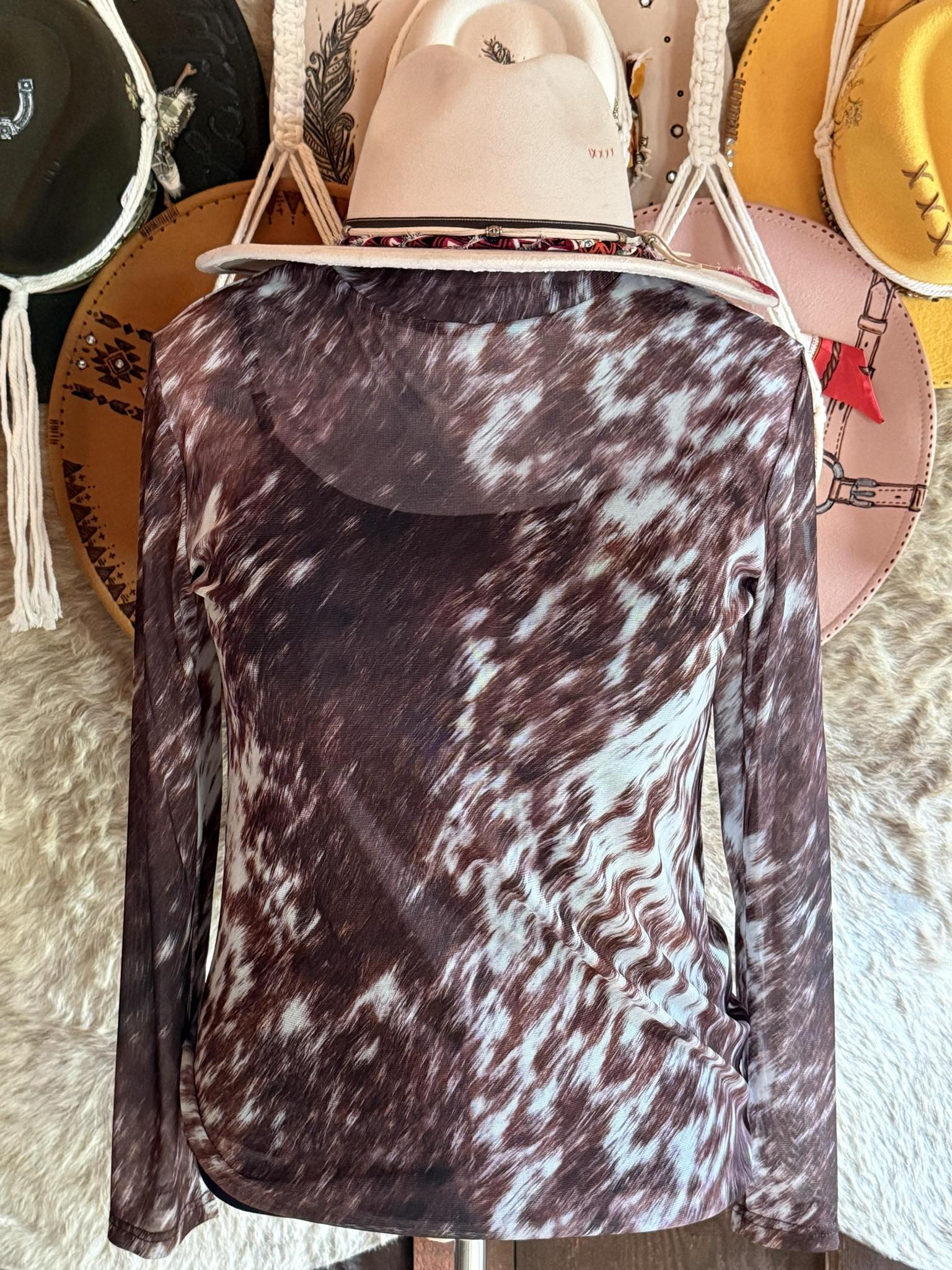 Brown and white cow-print mesh long-sleeve top displayed on a mannequin with a cream western hat, styled against a rustic background.