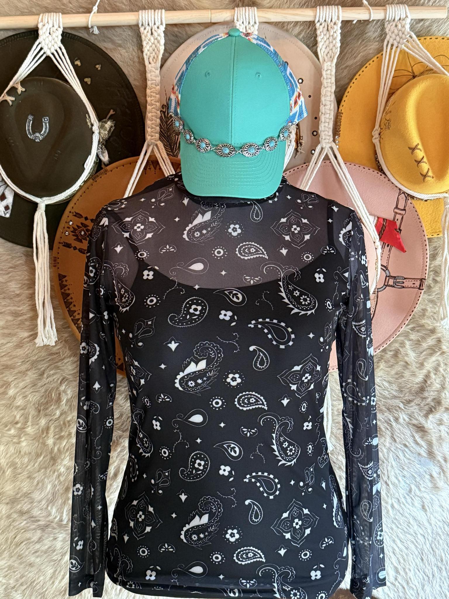 Black long-sleeve mesh top with white western paisley designs displayed on a mannequin wearing a turquoise cap and silver concho band.