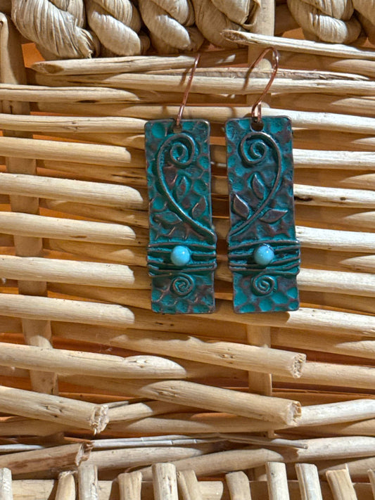 A pair of rustic copper rectangle earrings with embossed leaf and swirl designs, turquoise patina finish, and turquoise bead accent wrapped in coordinating wire, displayed on a woven wicker background.